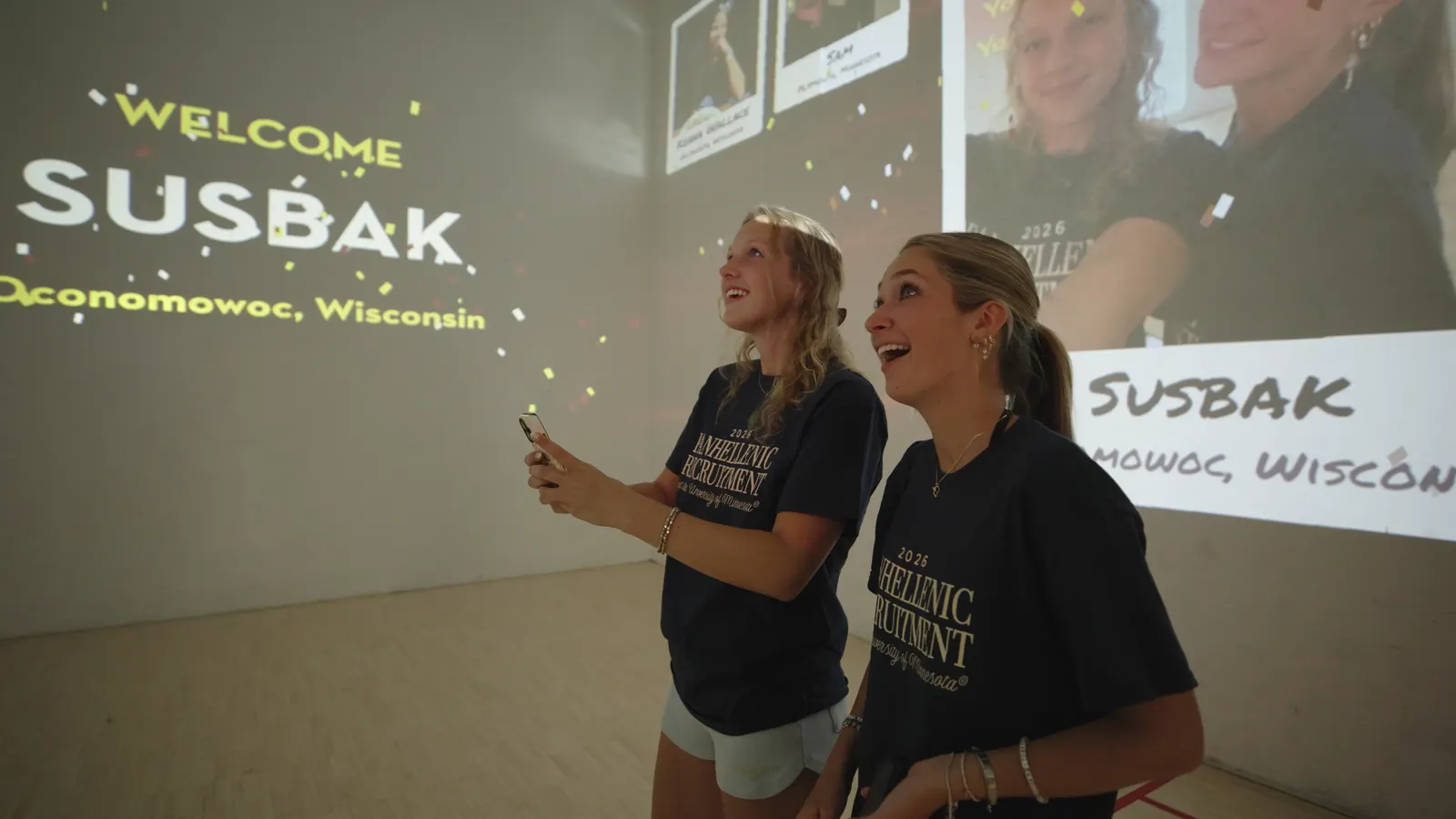 Two students laughing as a welcome for Oconomowoc, Wisconsin fills the wall behind them.