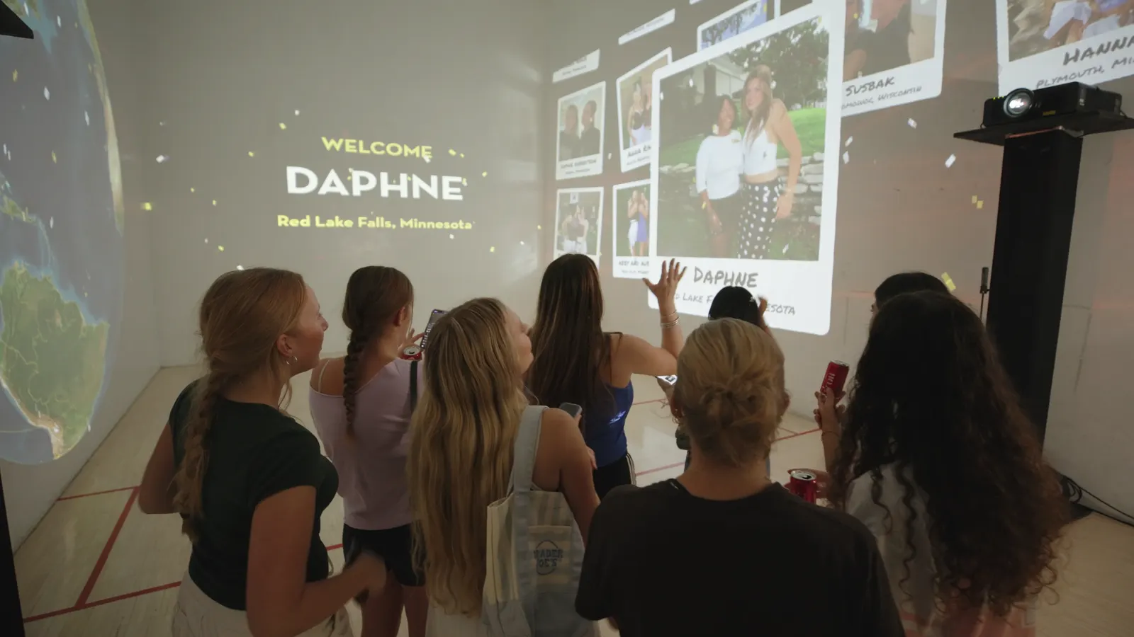 A group of students waving at a wall that reads “Welcome Daphne, Red Lake Falls, Minnesota” beside her photo.